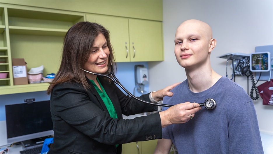 Dr. Katherine Janeway using a stethoscope to examine a patient's chest. The patient is bald and looking directly at the camera.