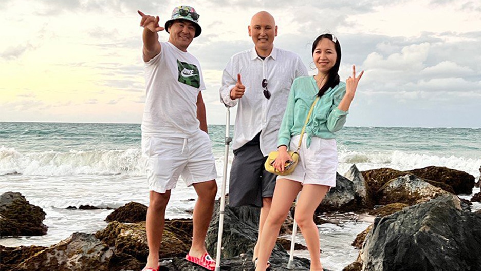 Osteosarcoma patient Talgat center standing on rocks at a beach, along with a friend and his girlfriend. They are all looking into the camera and smiling.