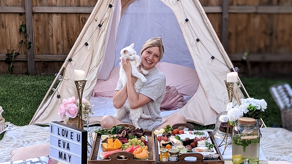 Osteosarcoma patient Eva sitting in a white triangular tent, with a tray of flowers and food spread out in front of her. She is holding a white cat and smiling.