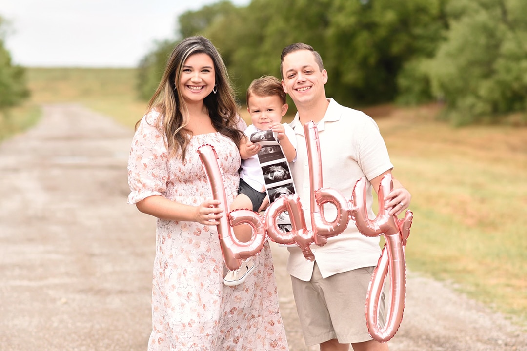 Shelbie, Kaleb Collins, and their son Graham holding an ultrasound printout and a balloon that says "baby"