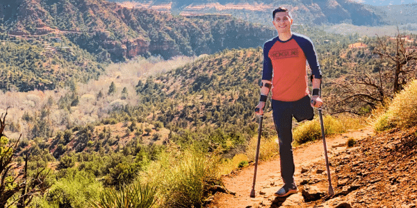 Carlos Garcia, two-time osteosarcoma survivor, is pictured on a hiking trail with gorgeous natural scenery behind him. He is using a cane on both arms, and you can see that his left leg has been amputated right below the hip.