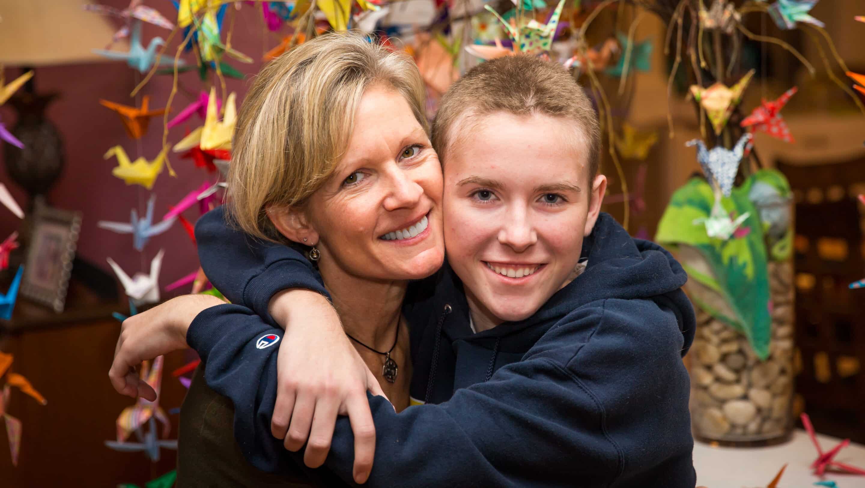 Gillian Vaughan (right) and her mom Connie hugging and smiling in front of the origami crane tree that friends and family created for her.