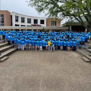 Large crowd of people wearing blue shirts with backs to the camera and one person in a yellow shirt in the middle of the front row