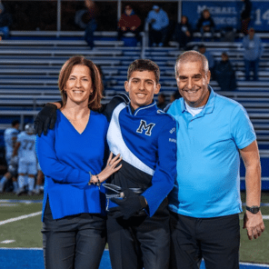 Gavin in his marching band uniform posing with his parents on a football field