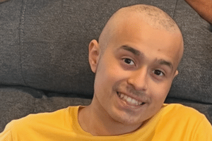 Dylan with a shaved head wearing a yellow shirt and smiling