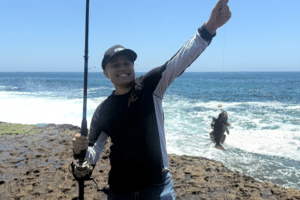 Dylan standing on the beach, holding a fishing rod in one hand and line with a fish on the end in the other