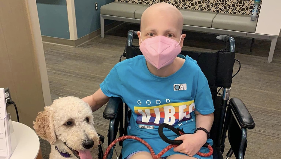 Grace sitting in her wheelchair with a mask on petting her dog.